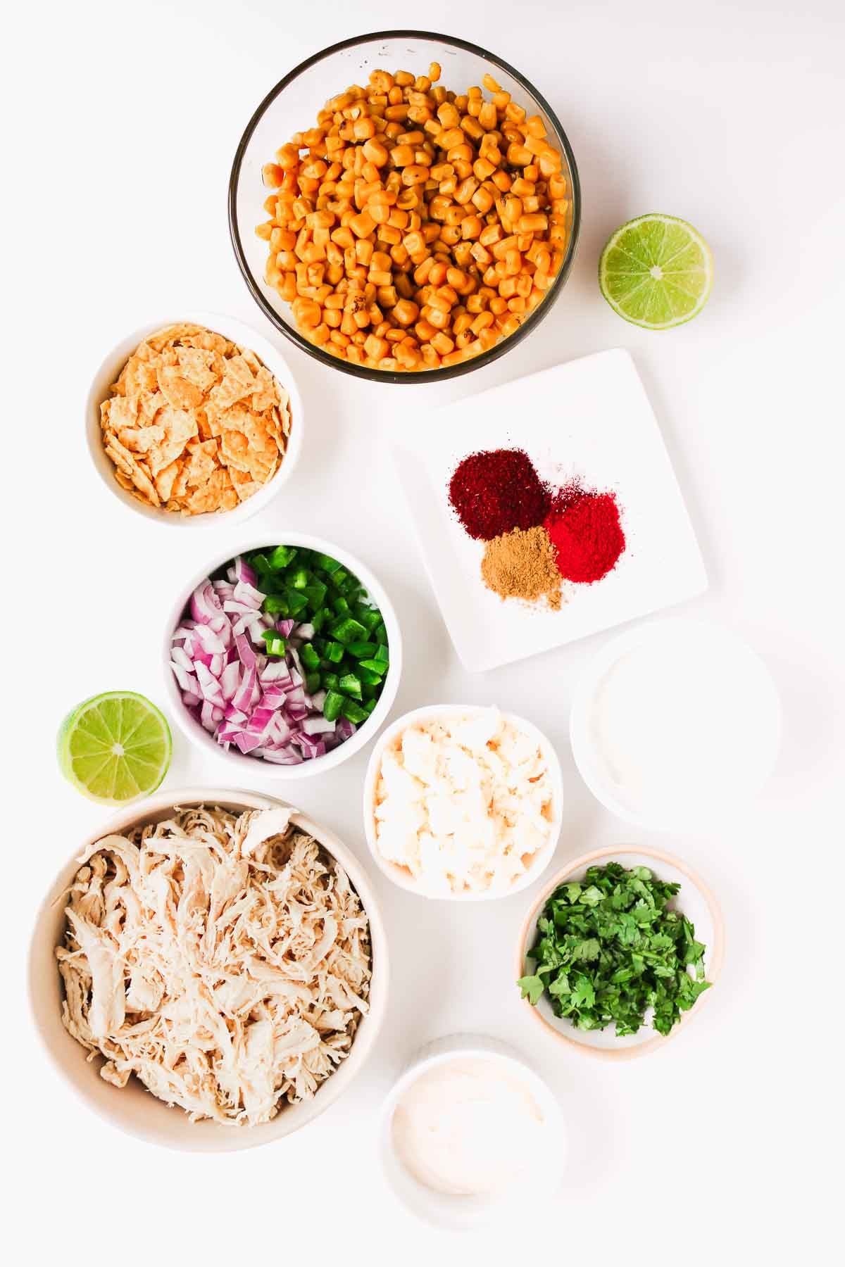 Bowls of corn, shredded chicken, onions, cilantro, cheese, spices, jalape&ntilde;os, and lime on a white background.