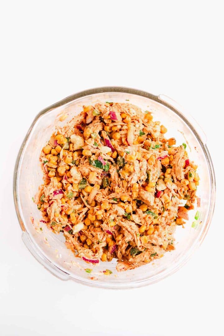 A glass bowl filled with a mixed chickpea salad with red onion, herbs, and shredded chicken on a white background.