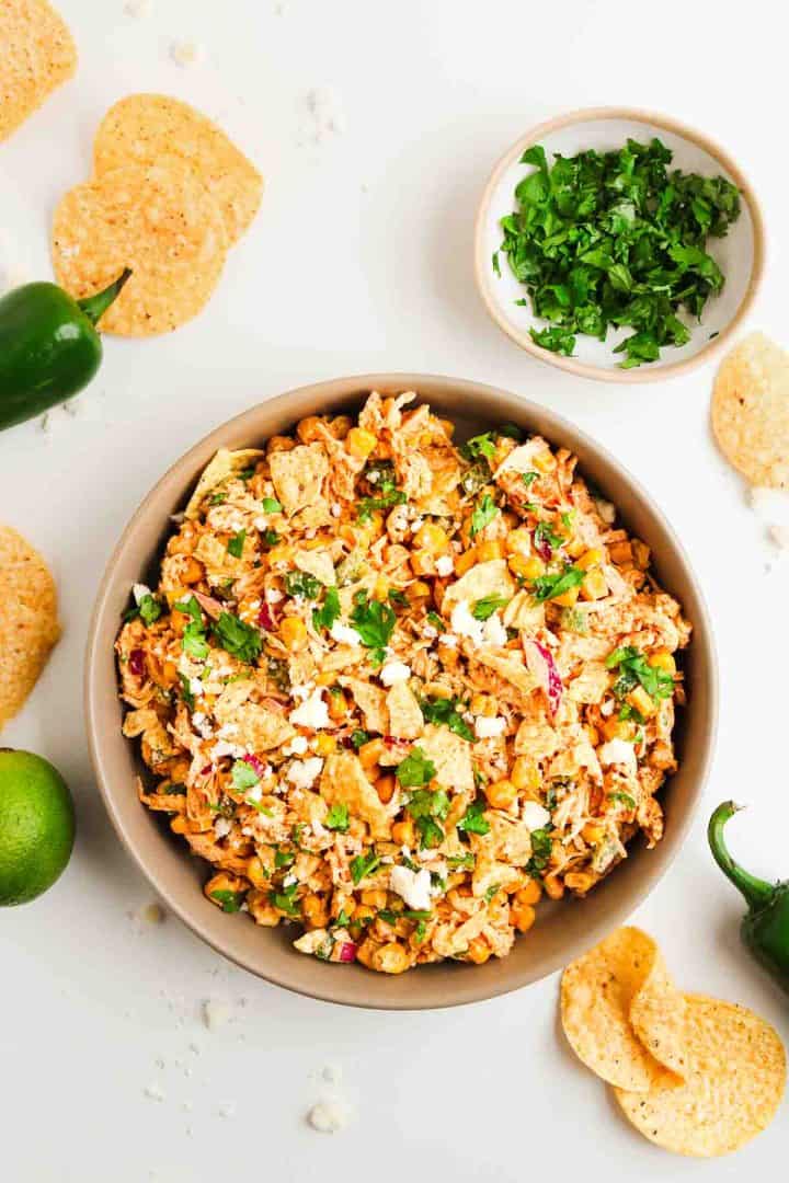 A bowl of Mexican street corn salad topped with cilantro, surrounded by tortilla chips, jalape&ntilde;os, and lime.