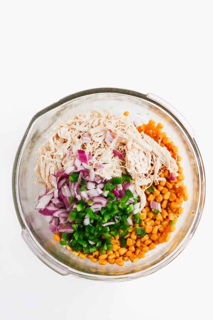 Glass bowl with shredded chicken, corn, diced red onion, and chopped green pepper on a white background.