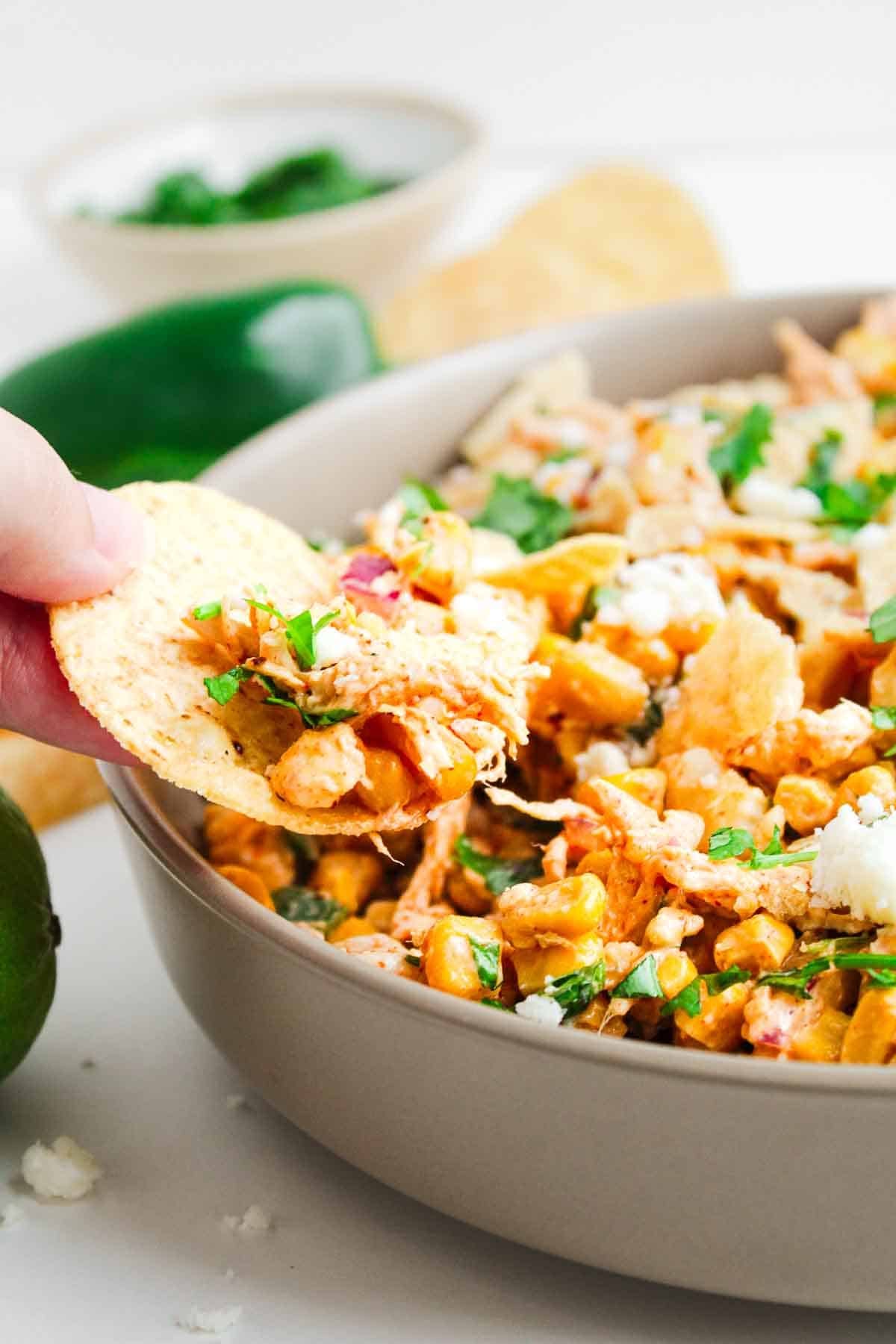 A hand scoops a cheesy bean dip with a tortilla chip from a bowl, garnished with fresh herbs.