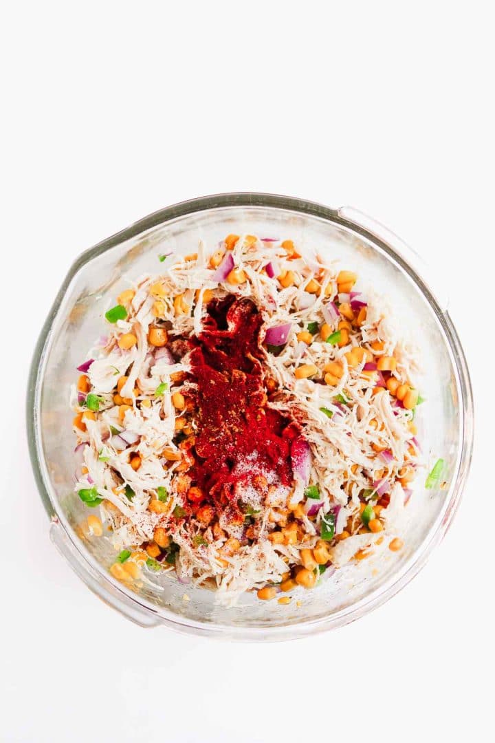 Glass bowl with shredded chicken, chickpeas, chopped onions, herbs, and spices on a white background.