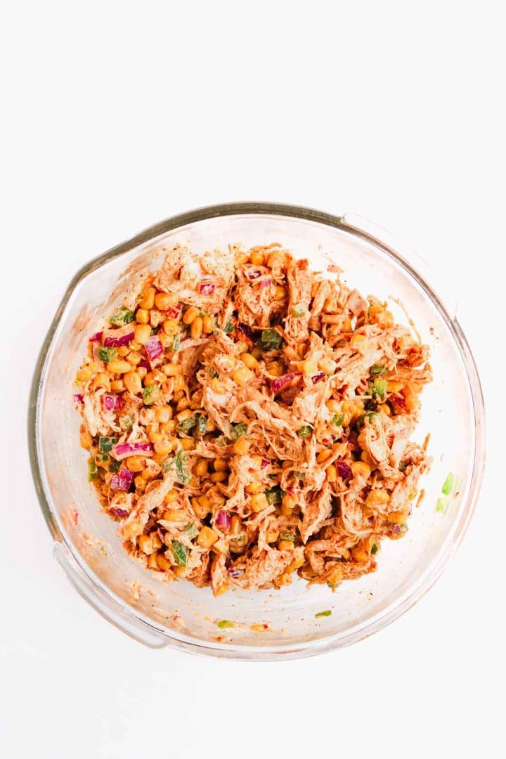 Glass bowl filled with shredded jackfruit, corn, red onions, and herbs mixed together on a white background.