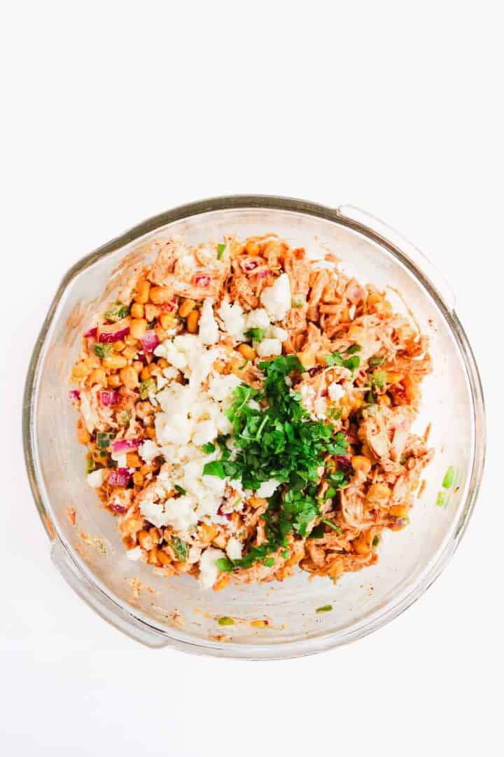 Glass bowl with shredded chicken, corn, red onion, cheese, and chopped cilantro on a white background.