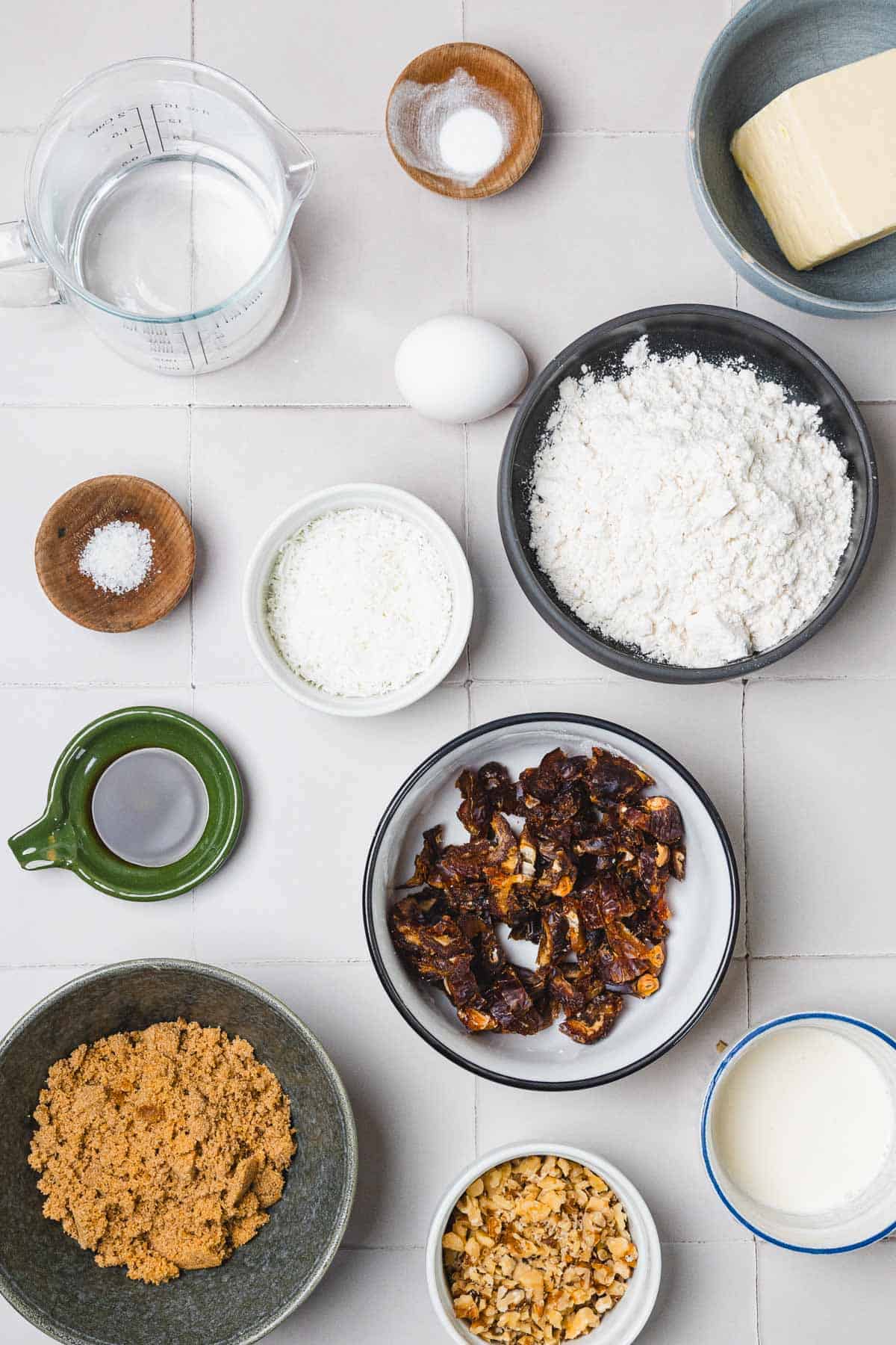 Top view of baking ingredients in bowls: flour, dates, walnuts, brown sugar, butter, egg, salt, water, and shredded coconut.