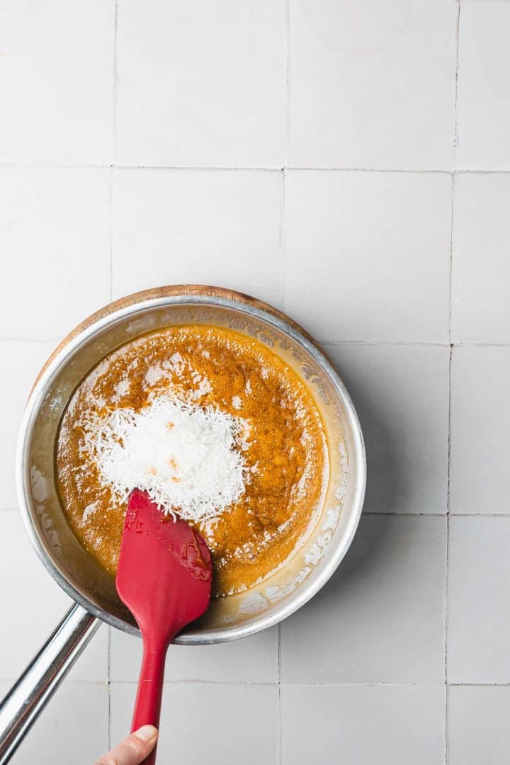 A hand stirs shredded coconut into a brown mixture in a saucepan using a red spatula.
