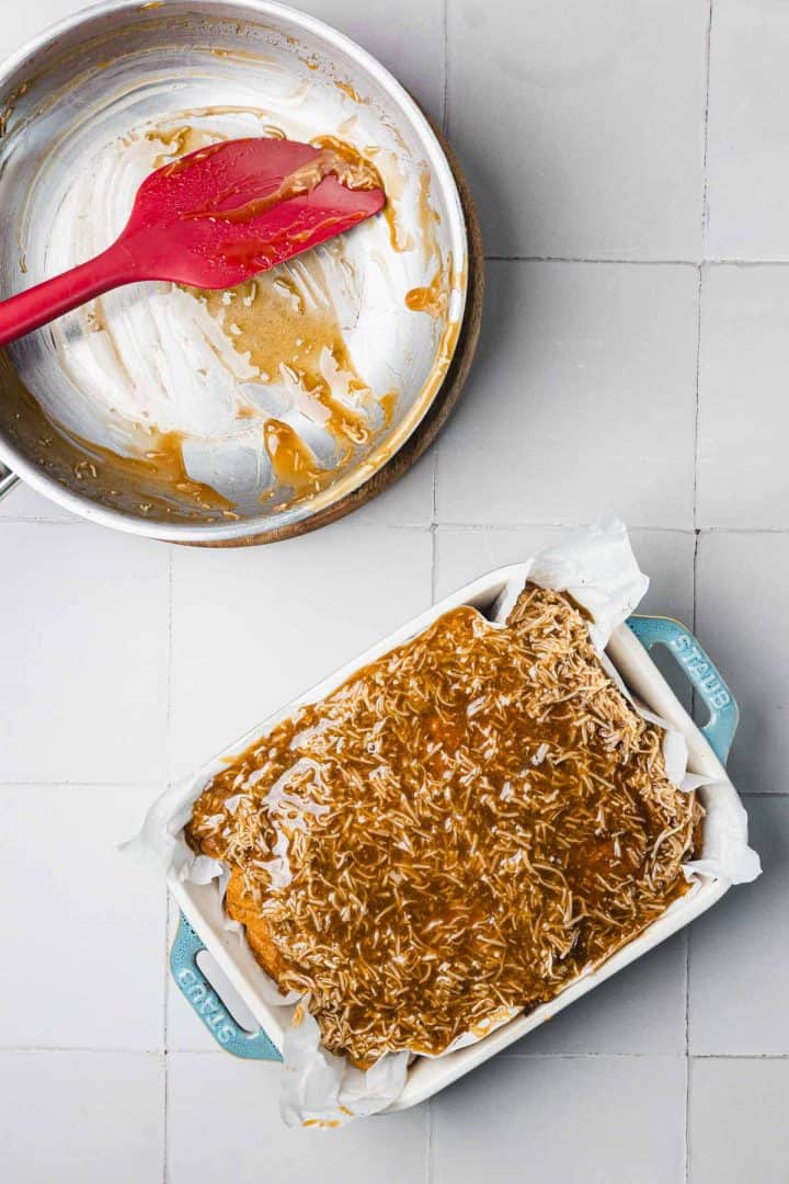 A baking dish with oat bars and caramel, next to a saucepan with caramel residue and a red spatula.