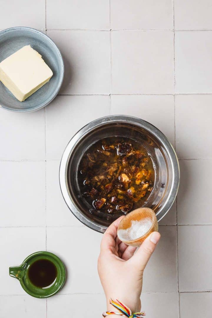 A hand adds salt to a bowl of dates in liquid, with butter and vanilla on a tiled surface nearby.