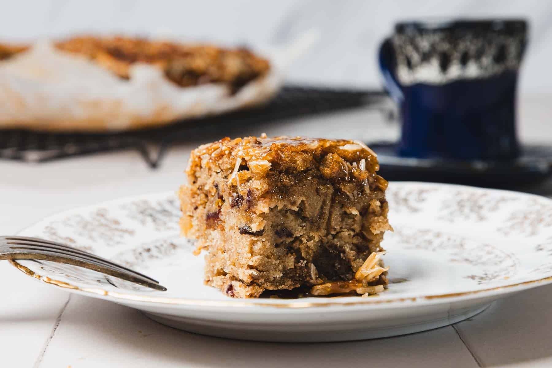 A square slice of cake on a decorative plate with a fork, blurred cake and cup in the background.