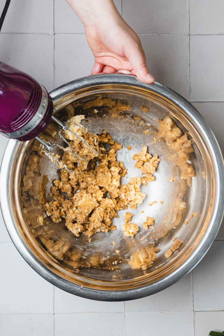 A hand holds a mixer blending cookie dough in a large metal bowl on a tiled surface.