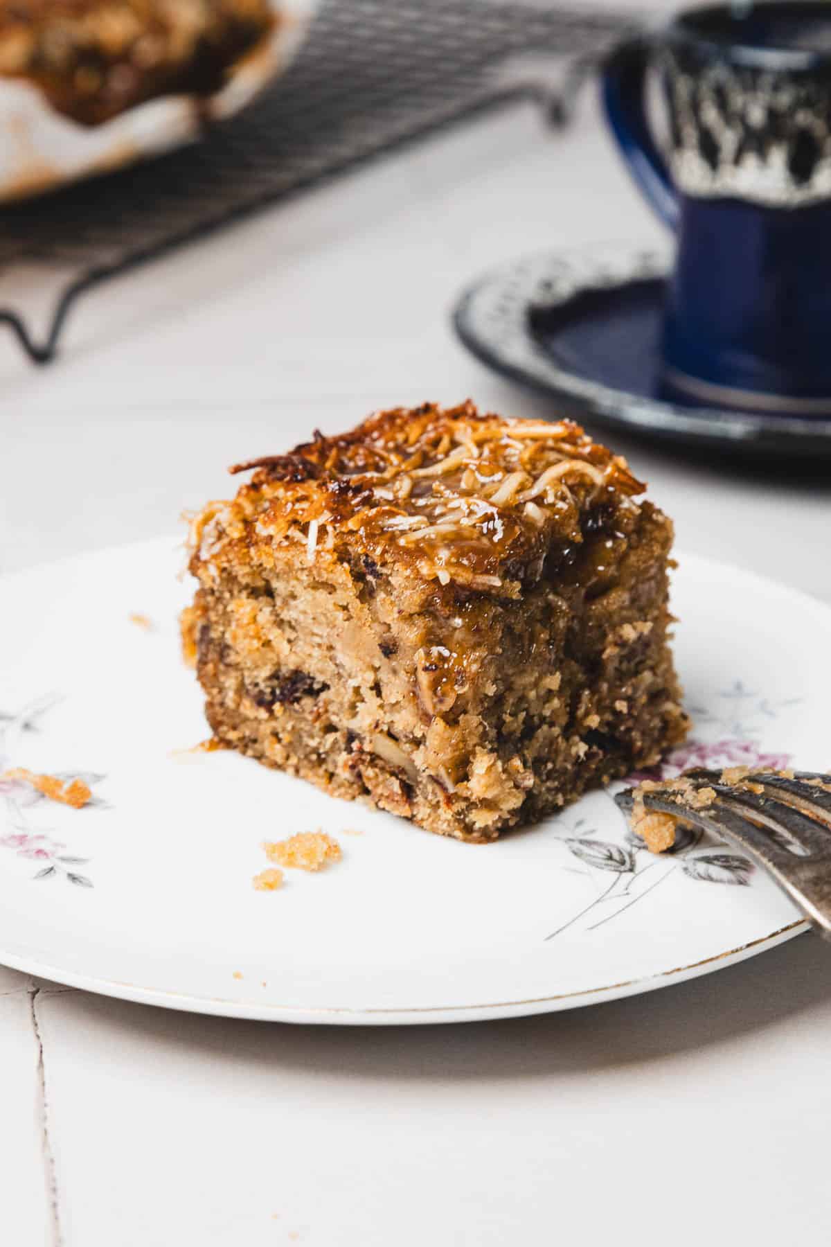 A slice of moist Queen Elizabeth Cake with a caramelized topping sits on a floral plate, accompanied by a fork and a cup in the background.