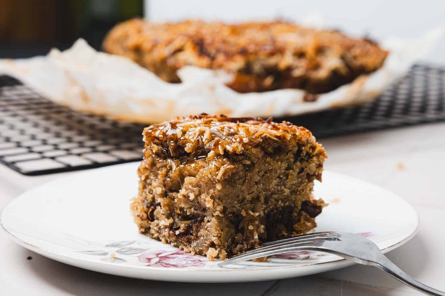 A slice of moist cake with raisins sits on a plate, with the rest of the cake in the background.