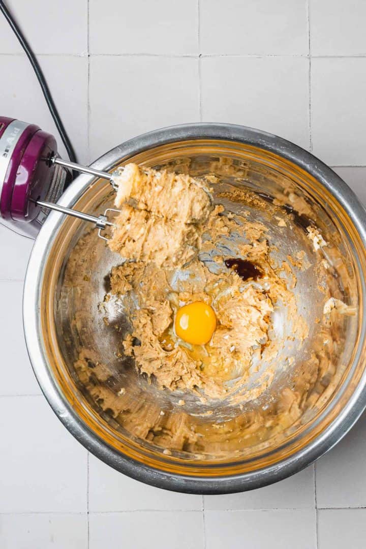A mixing bowl with cookie dough, an egg, and a hand mixer on a tiled countertop.