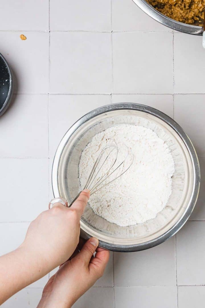 A hand whisking flour in a metal bowl on a tiled countertop, with baking ingredients nearby.