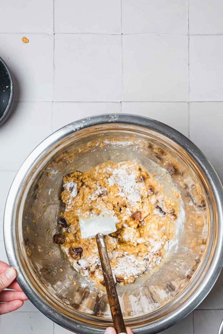 Hands mixing cookie dough with chocolate chips in a large stainless steel bowl using a spatula on a tiled surface.