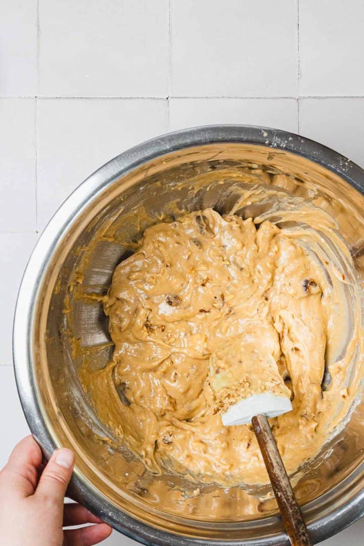A hand holds a bowl of cookie dough with a spatula resting inside, on a tiled surface.