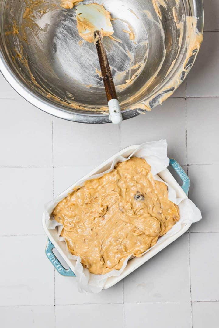 A loaf pan with unbaked batter on parchment paper beside a mixing bowl with a spatula.