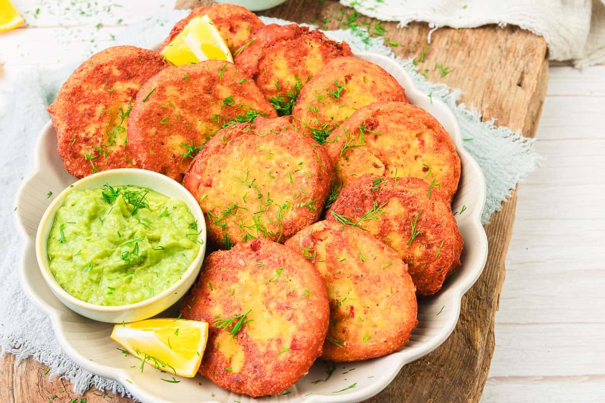A plate of golden-brown red lentil patties with lemon wedges and a small bowl of green dip on a rustic wooden surface.