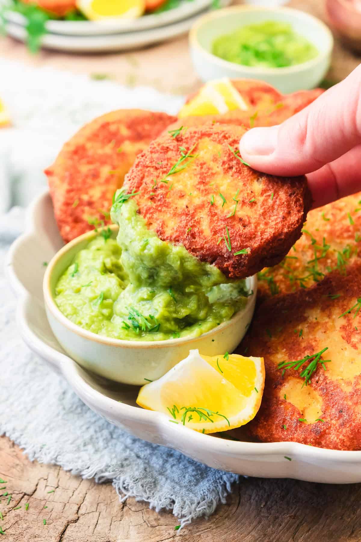 A hand dips a crispy red lentil patty into a bowl of green sauce, with lemon wedges and more red lentil patties on the plate.