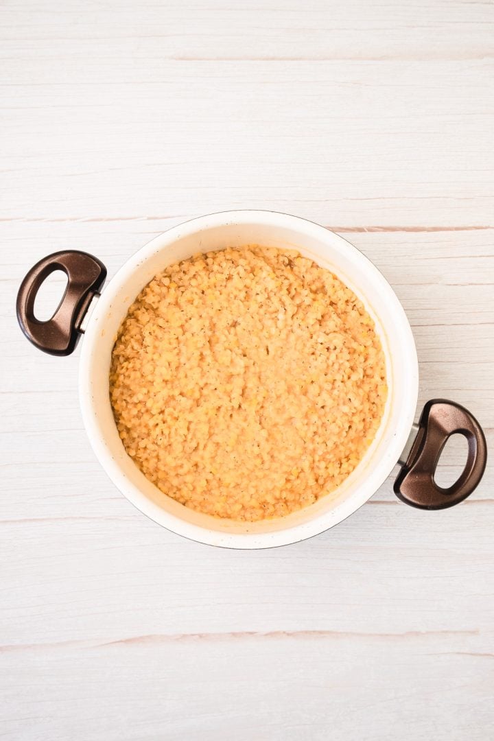 A white pot with black handles filled with cooked red lentils, perfect for making delicious red lentil patties, sits on a light wooden surface.