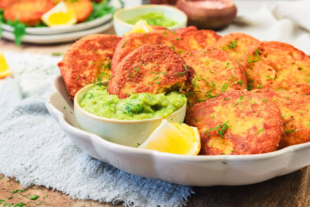 A plate of golden red lentil patties with a bowl of green dip and lemon wedges on a rustic cloth.
