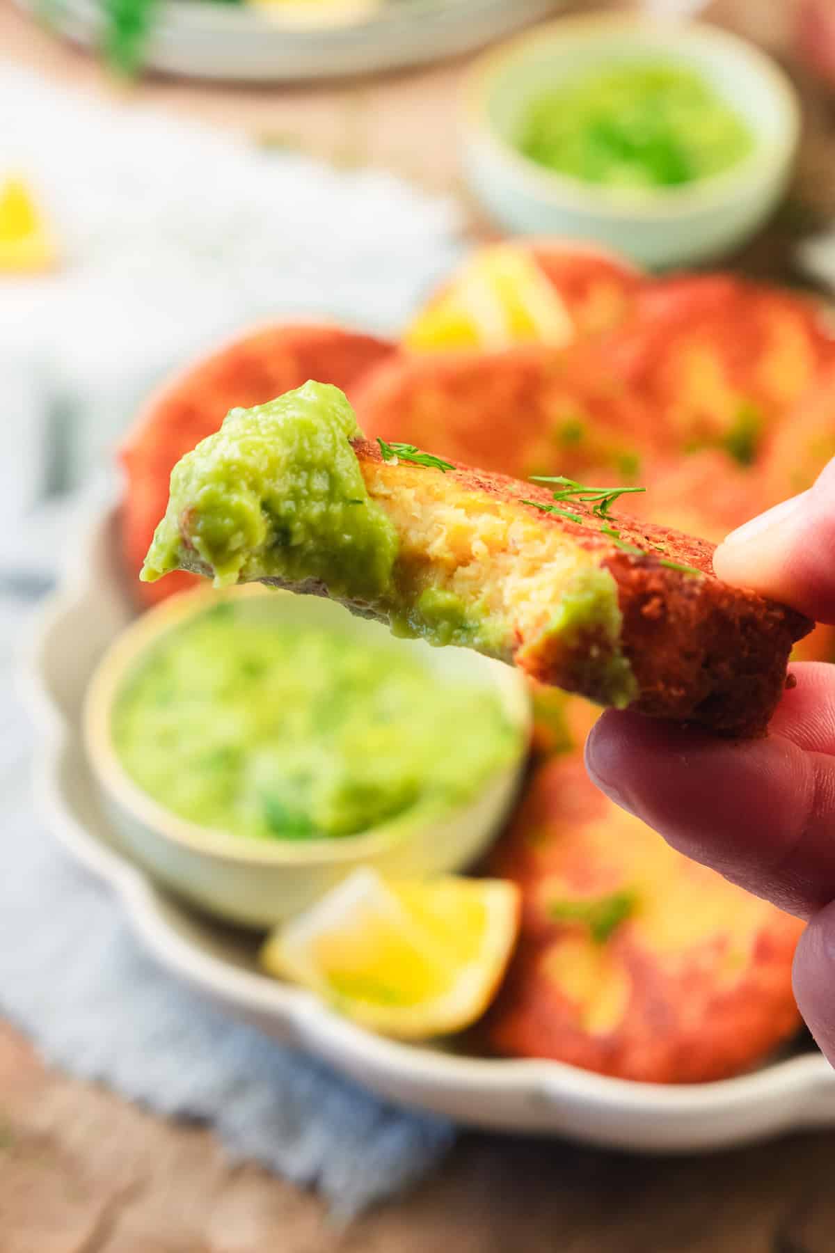 A hand holds a crispy red lentil patty fritter dipped in green sauce, with more red lentil patties and sauce in the background.