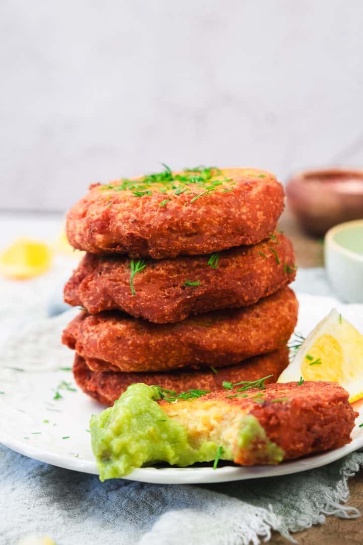 A stack of golden red lentil patties on a plate, garnished with herbs, with one patty dipped in green sauce.