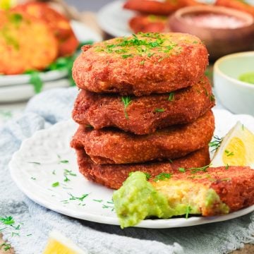 Four crispy fritters stacked on a plate with green dip, lemon wedges, and fresh herbs sprinkled on top.
