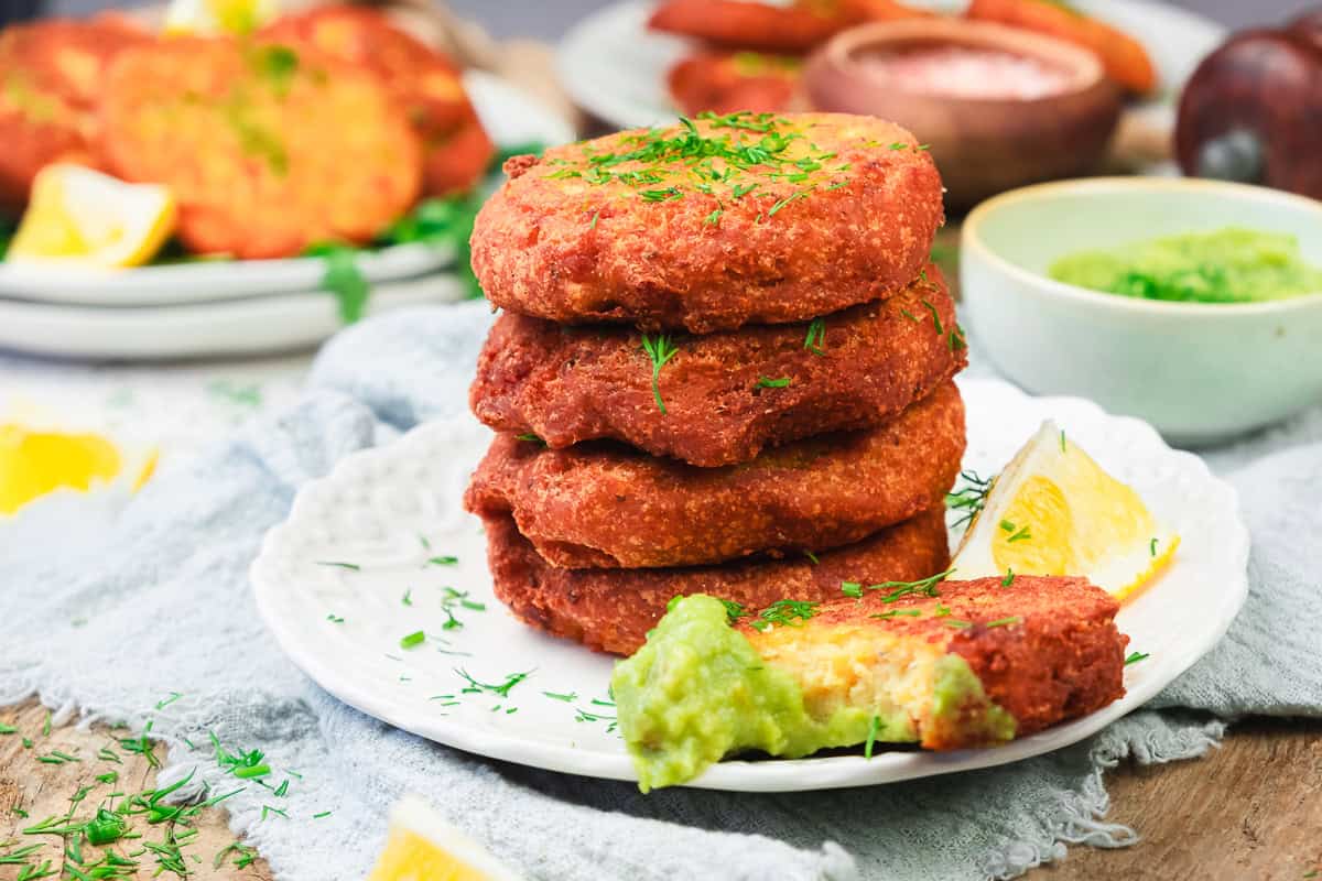 Four crispy fritters stacked on a plate with green dip, lemon wedges, and fresh herbs sprinkled on top.