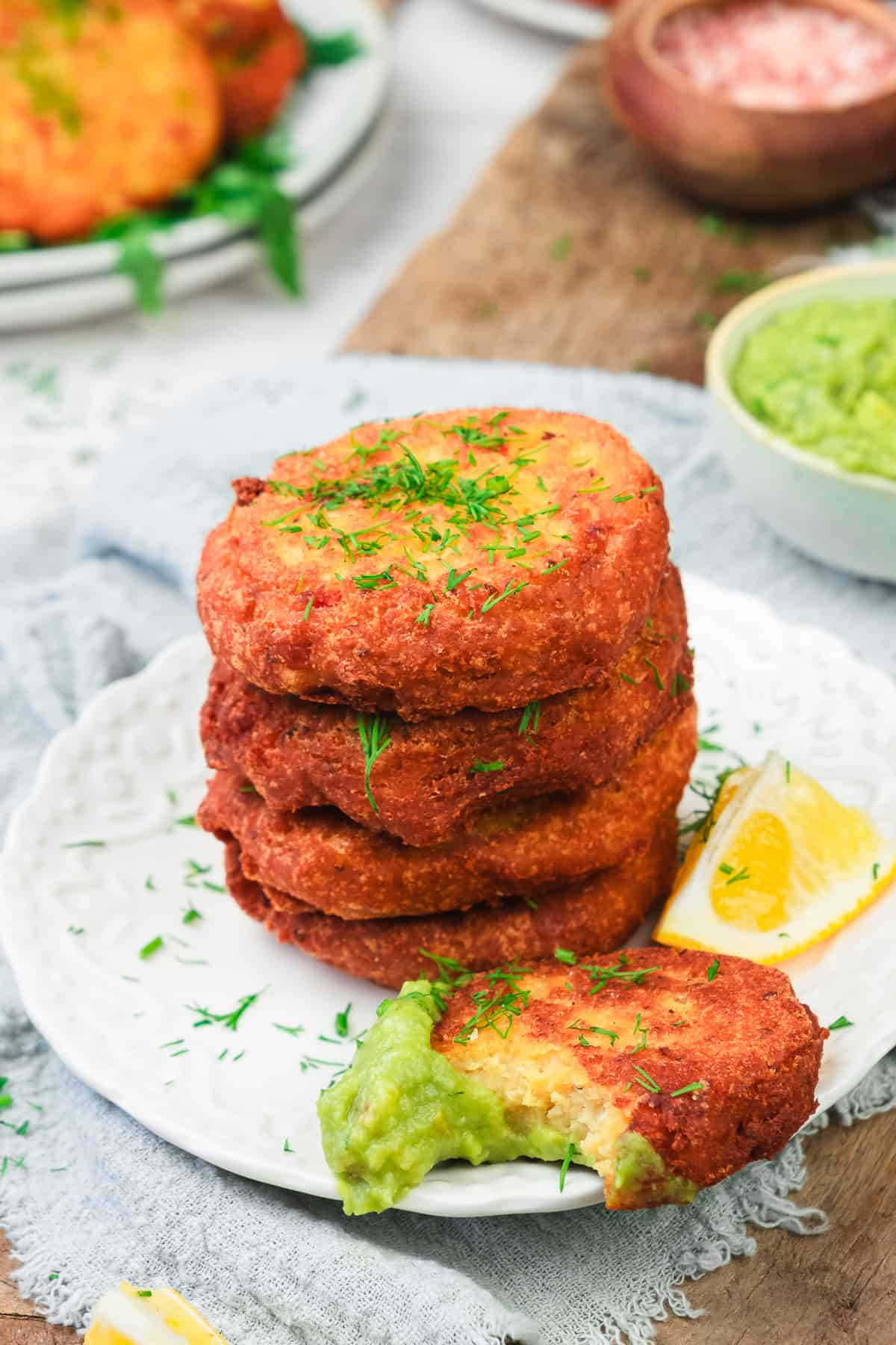 A stack of golden-brown fritters on a plate with herbs, lemon wedges, and green dipping sauce.