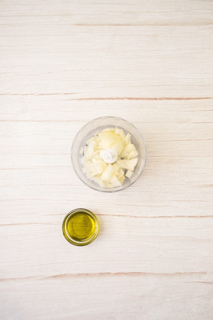 Chopped onions in a food processor and a small bowl of olive oil on a light wooden surface, perfect for preparing red lentil patties.