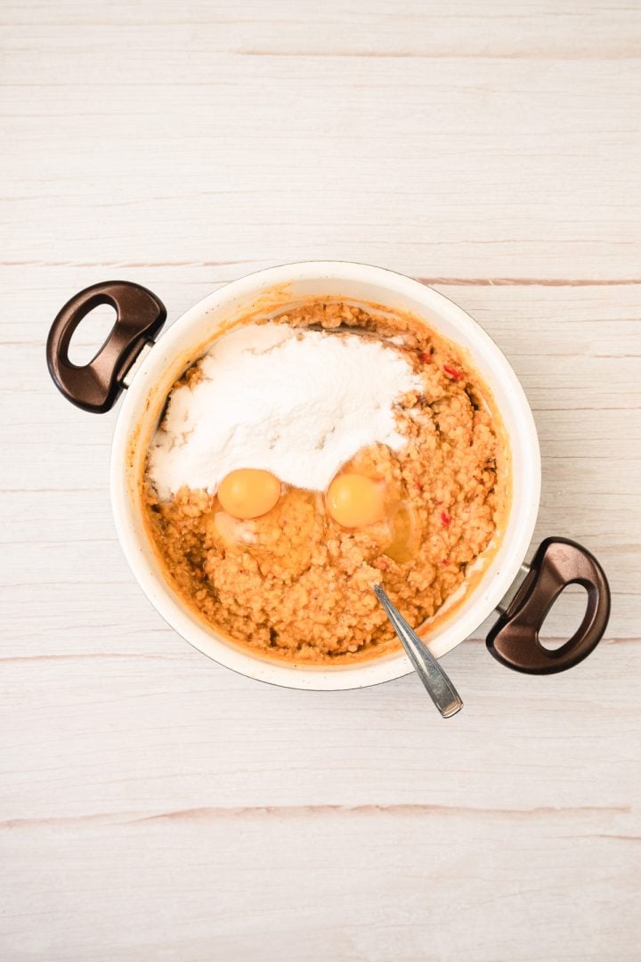 A mixing bowl with batter for red lentil patties, two cracked eggs, and flour, with a spoon resting inside.