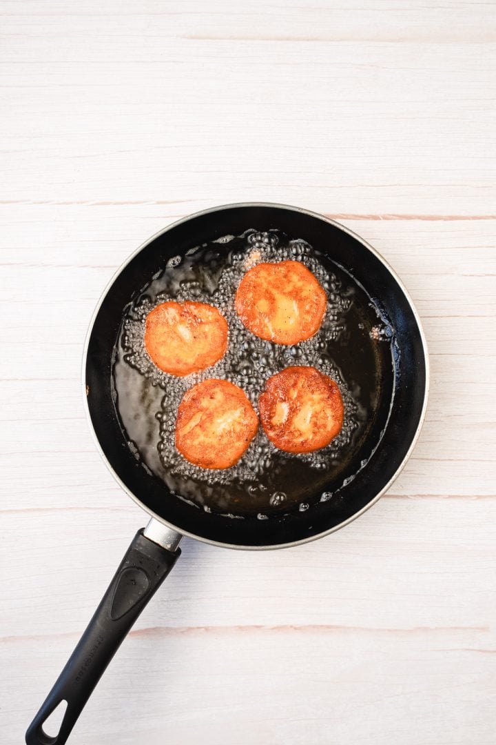 Four round red lentil patties frying in oil in a black pan on a light-colored wooden surface.