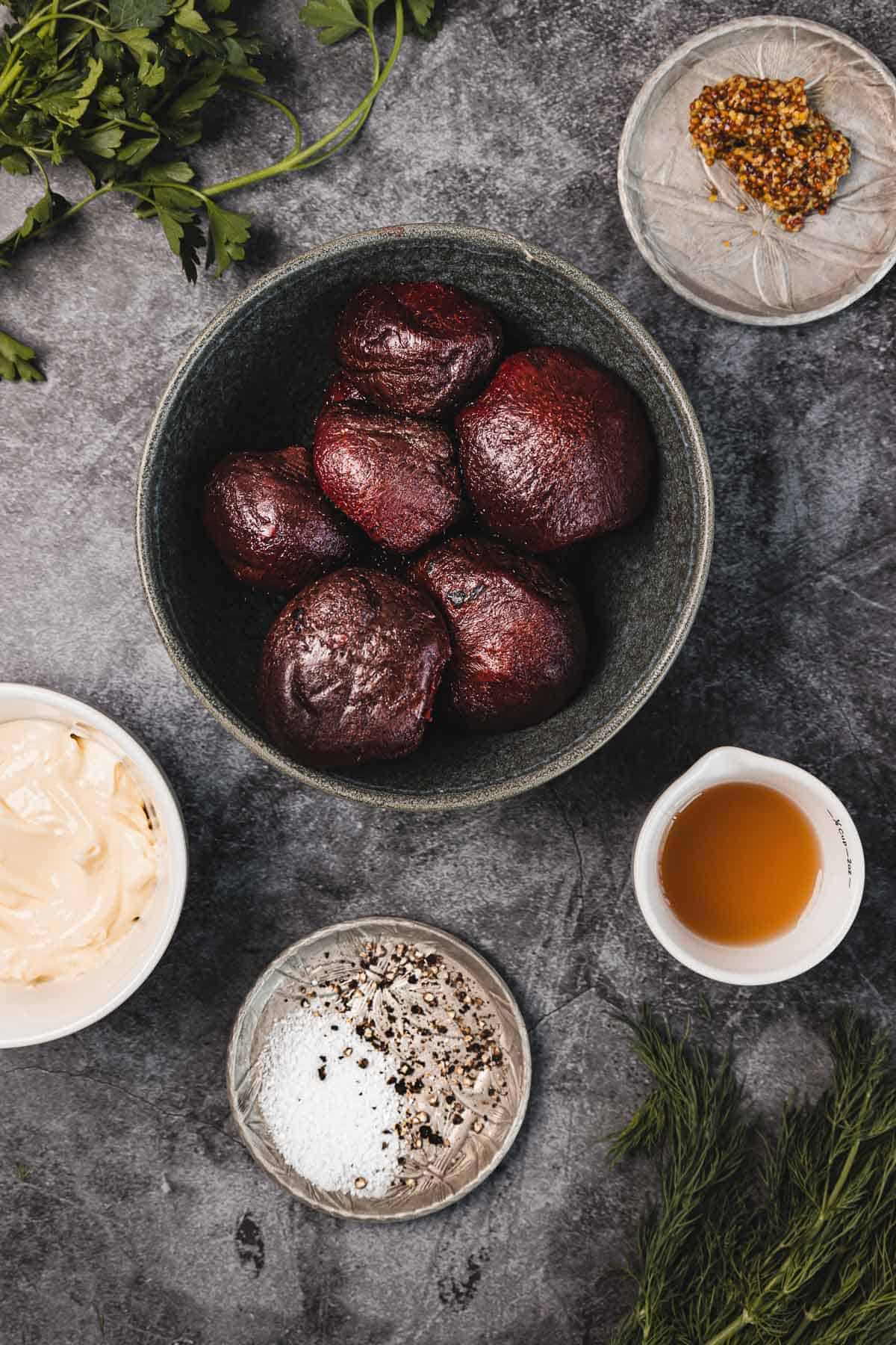 A bowl of roasted beets surrounded by mustard, mayonnaise, vinegar, salt, pepper, and fresh herbs on a gray surface.