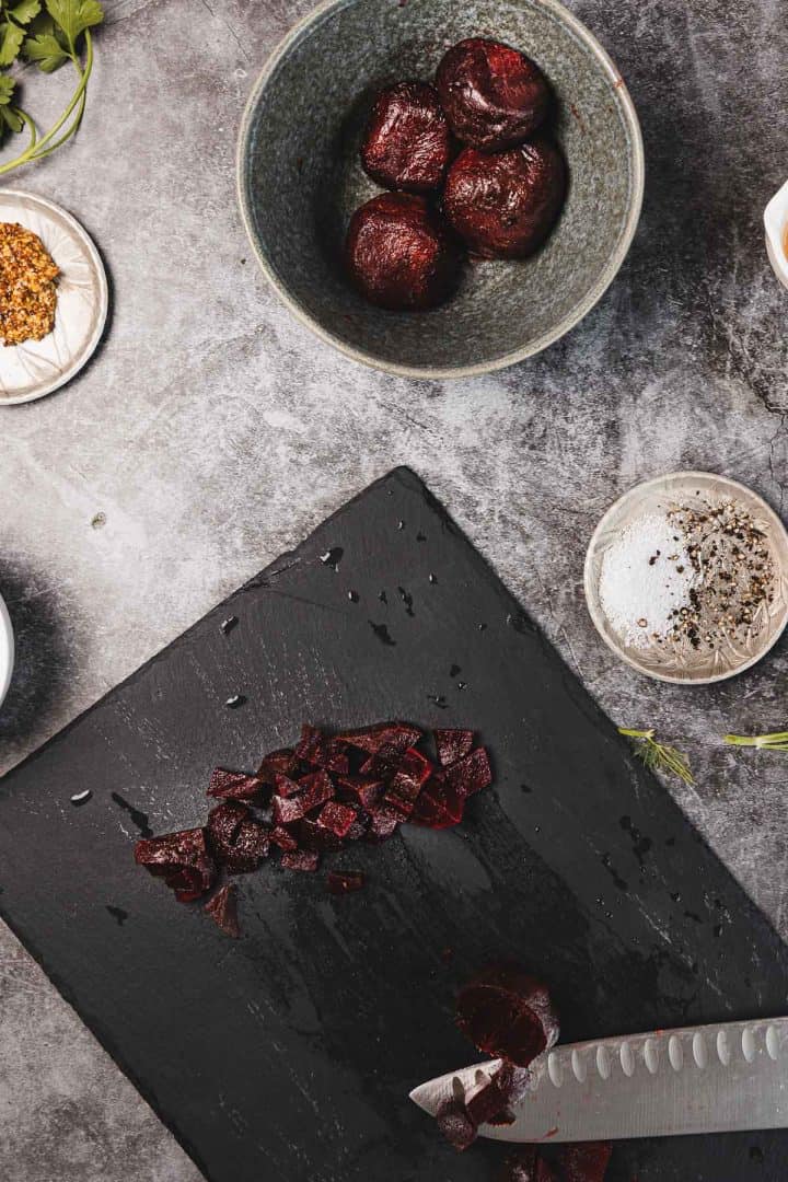 Chopped beets on a black cutting board with a knife, whole beets in a bowl, and spices nearby.