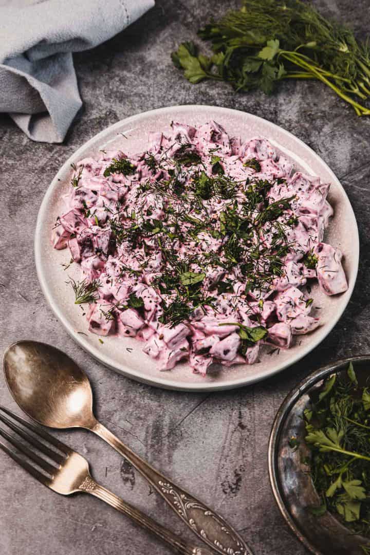 A plate of creamy beet salad garnished with fresh herbs, next to cutlery and a bunch of greens.