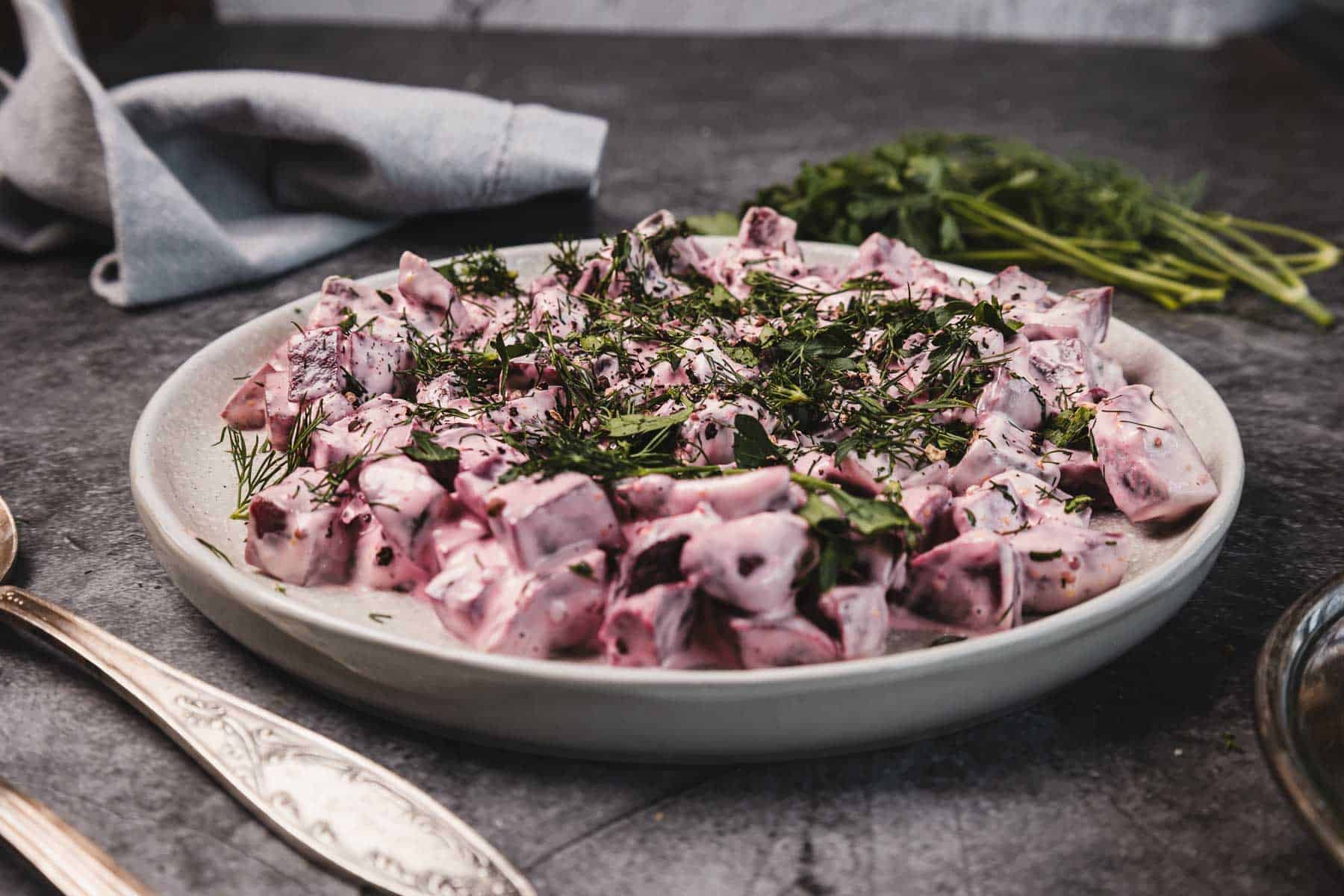 A plate of creamy beet salad garnished with fresh dill sits on a dark table next to silverware and herbs.