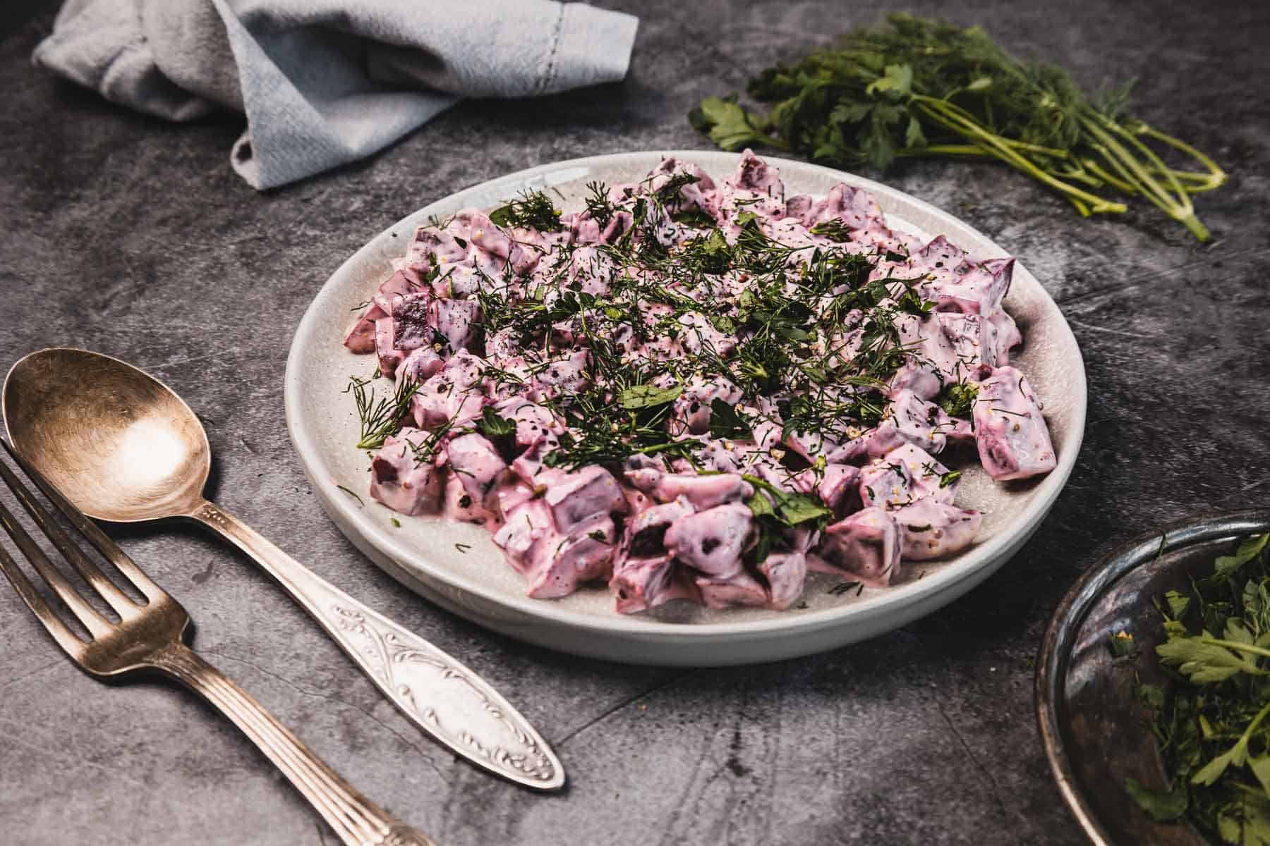 A plate of creamy beet salad garnished with fresh herbs, next to a fork and spoon on a dark surface.