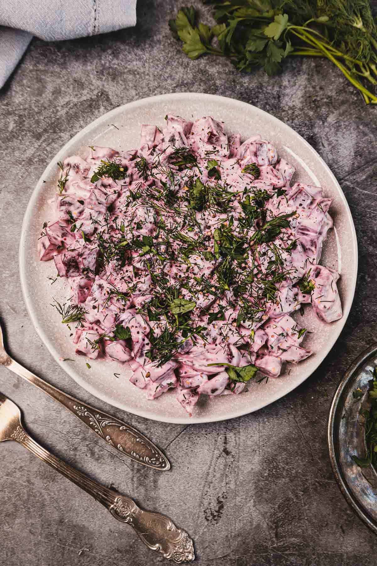 A plate of creamy beet salad garnished with chopped herbs, with antique silverware beside it on a gray surface.