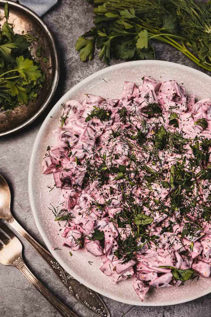 A plate of creamy beet salad topped with fresh herbs, with parsley and spoons beside it on a gray surface.