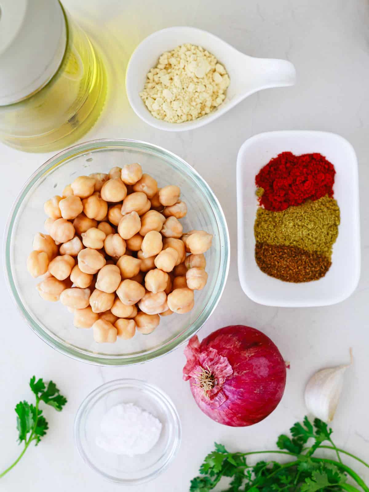 Chickpeas, spices, onion, garlic, oil, herbs, baking powder, and salt arranged on a white surface.