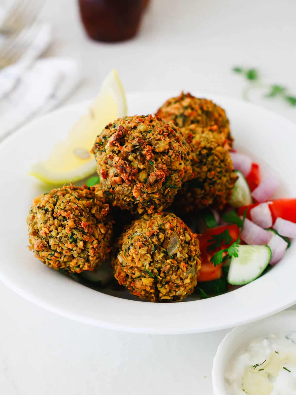 A bowl of falafel balls with sliced cucumber, tomato, red onion, and lemon wedges.