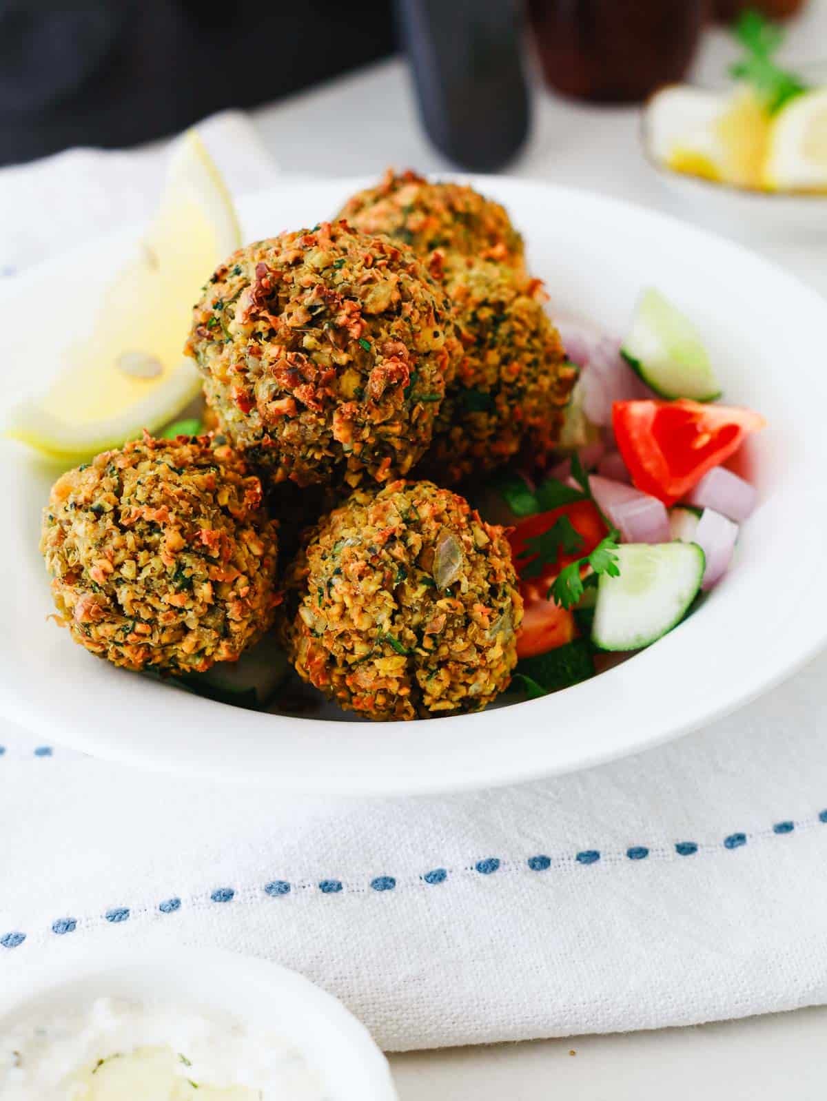 Four falafel balls with sliced cucumber, tomato, onion, and lemon wedges on a white plate.