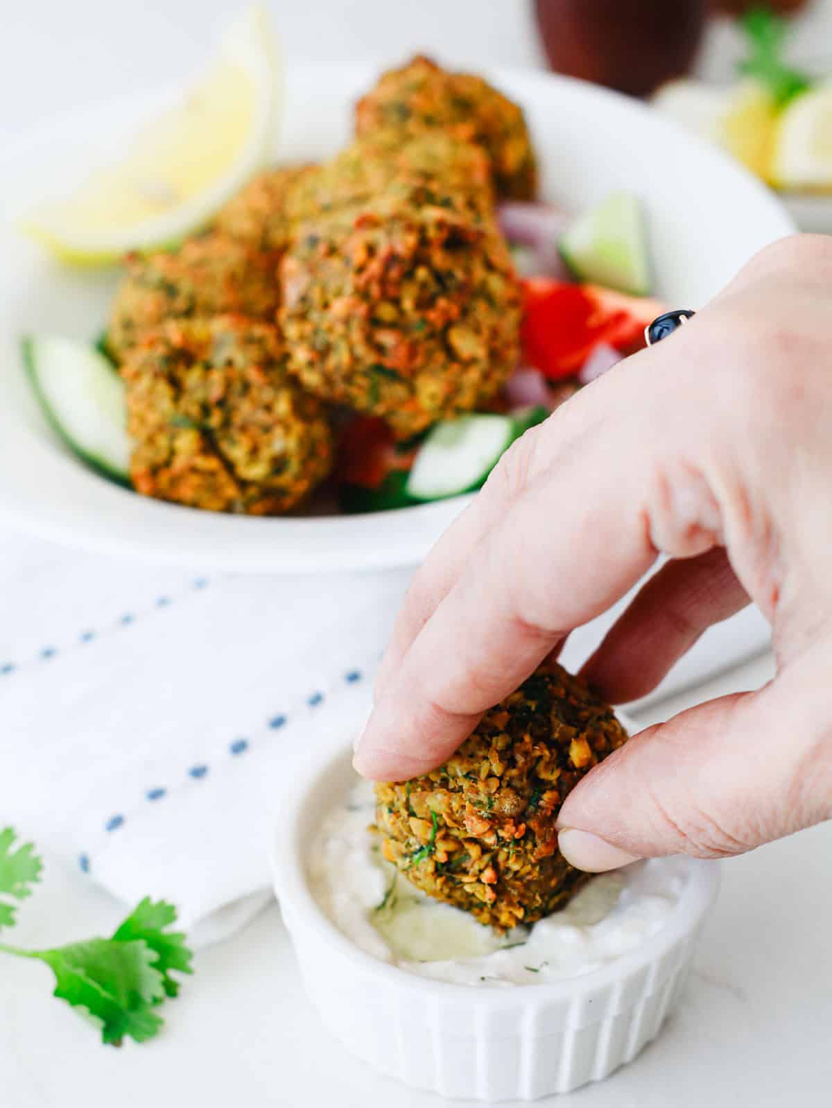 A hand dips a falafel ball into a white sauce, with more falafel and veggies in a bowl in the background.