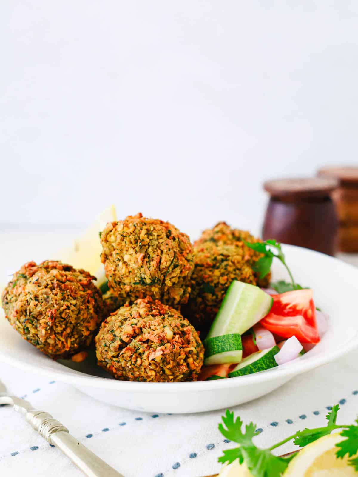 A white bowl of falafel balls served with fresh cucumber, tomato, onion, and a lemon wedge.