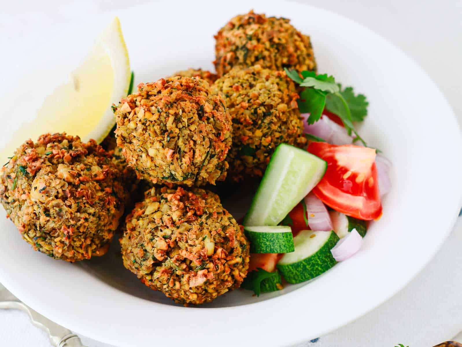 Five falafel balls with sliced cucumber, tomato, onion, and a lemon wedge on a white plate.