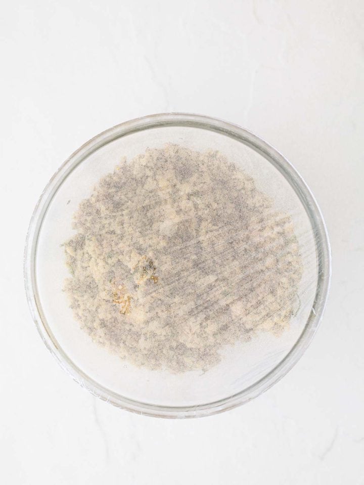 A glass bowl covered with plastic wrap, containing a crumbly mixture on a white surface.