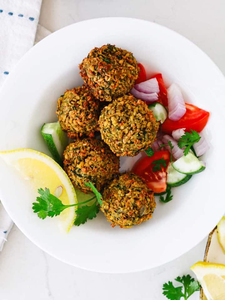 Five falafel balls served with chopped tomatoes, cucumbers, onions, a lemon wedge, and cilantro on a white plate.