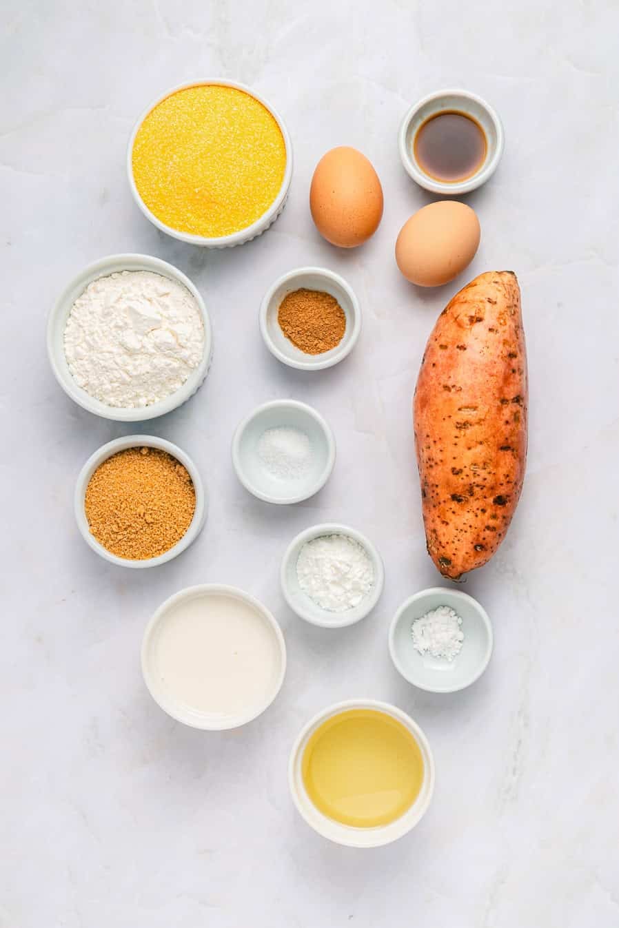 Various baking ingredients, including those for Sweet Potato Cornbread—such as a sweet potato, eggs, flour, and spices—are artfully arranged on a marble surface.