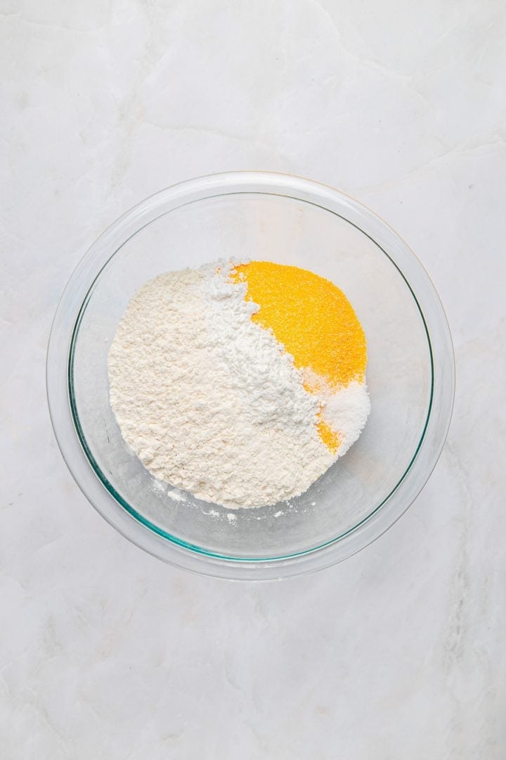 A glass bowl with flour, cornmeal, and baking powder on a white marble surface—perfect ingredients for making delicious Sweet Potato Cornbread.